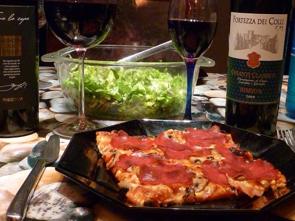 A pepperoni pizza on a crowded dinner table with wine bottles, photographed on a phone